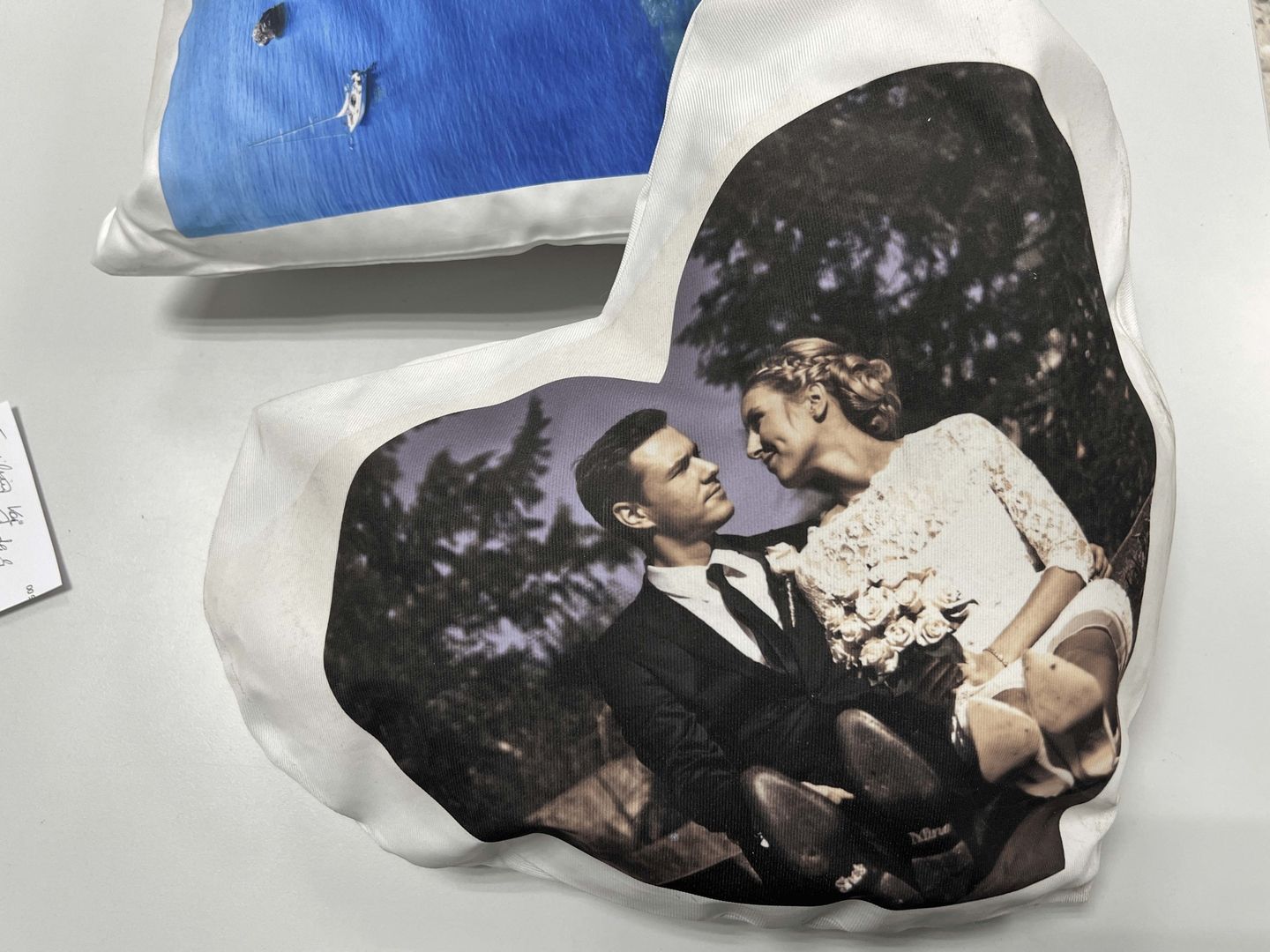 Ein herzförmiges Kissen, bedruckt mit einem Hochzeitsfoto. Das Foto zeigt ein Hochzeitspaar in festlicher Kleidung, das sich lächelnd ansieht. Der Hintergrund des Kissenaufdrucks ist dezent, um das Bild hervorzuheben. Das Kissen liegt auf einer glatten Oberfläche.